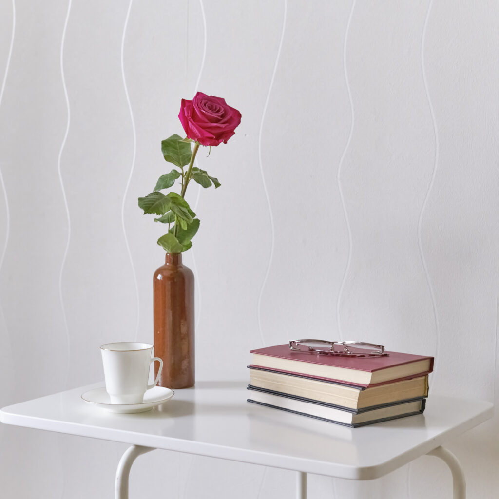 Composition reference image depicting a teacup, a rose in a small vase, and a pile of books with a pair of glasses on top of a modern white table.